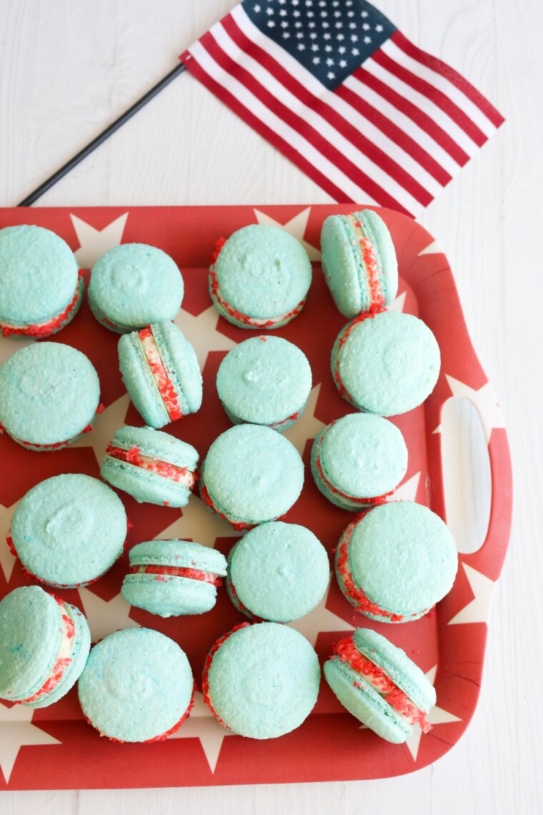 Blue rocket pop macarons with red and white filling chill on a tray beside a mini American flag on a plain white surface.