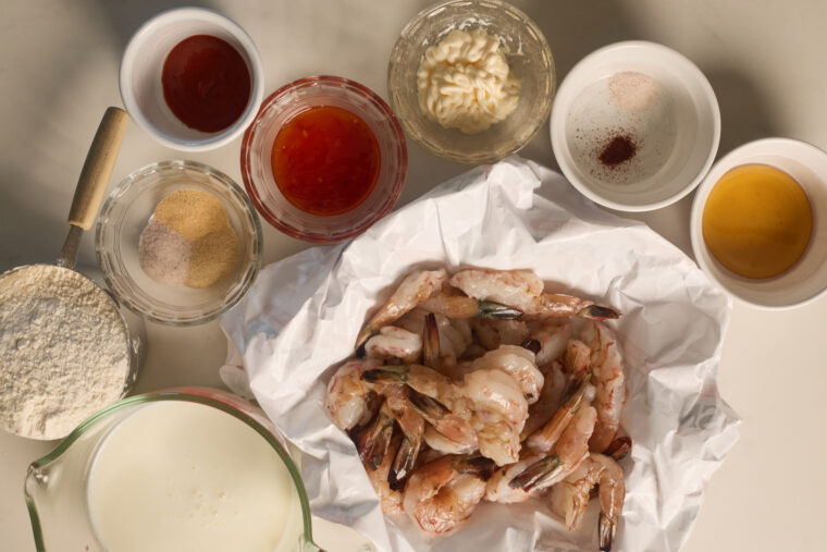 Raw shrimp ready for bang bang shrimp, with bowls of flour, spices, sauces, oil, and milk sitting on a light surface.