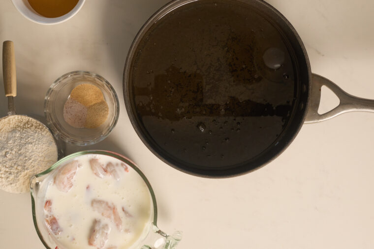 Top-down shot: pan with oil, seasoned flour, bang bang shrimp spice, buttermilk-soaked chicken, and a liquid bowl on a light counter.