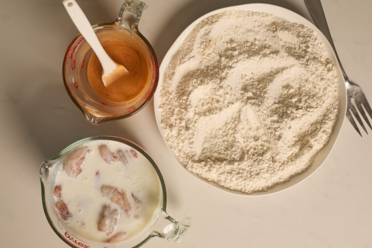 Three bowls with flour, a cup of seasoned liquid, and chicken pieces soaking in buttermilk for bang bang shrimp.