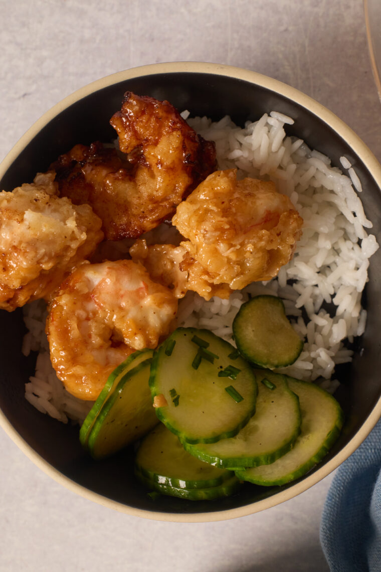 A bowl with fluffy white rice, crispy bang bang shrimp, and fresh cucumber salad tossed with herbs.
