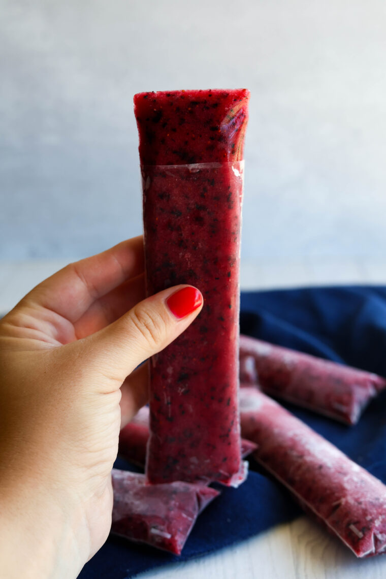 A hand with red nails grabs a homemade berry ice pop in a plastic sleeve, more pops scattered on a dark cloth behind it.