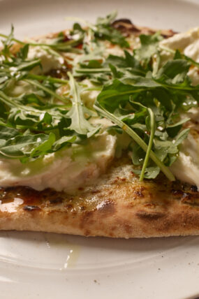 A slice of burrata pizza loaded with gooey cheese and fresh arugula sits on a white plate, close-up.