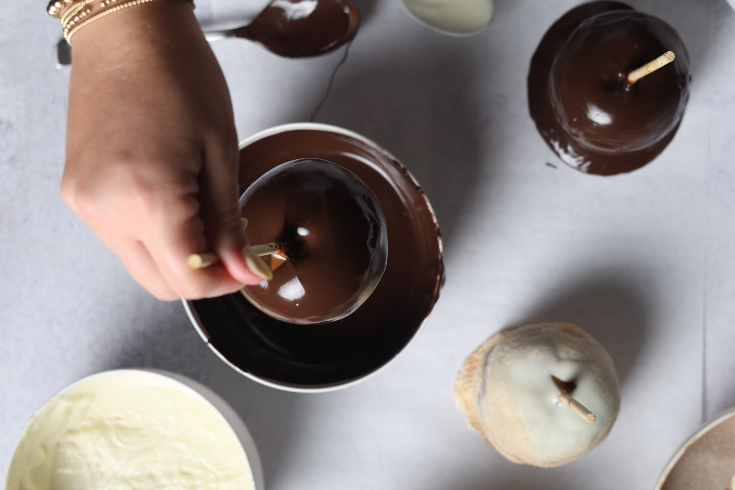 Someone&rsquo;s dipping an apple on a stick into gooey chocolate, with bowls of white chocolate, chips, and toppings all around.