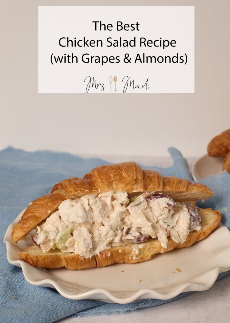 A croissant stuffed with chicken salad, grapes, and almonds sits on a scalloped plate with a blue cloth in the background.