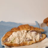 A croissant stuffed with chicken salad, grapes, and celery rests on a white plate over a blue cloth.