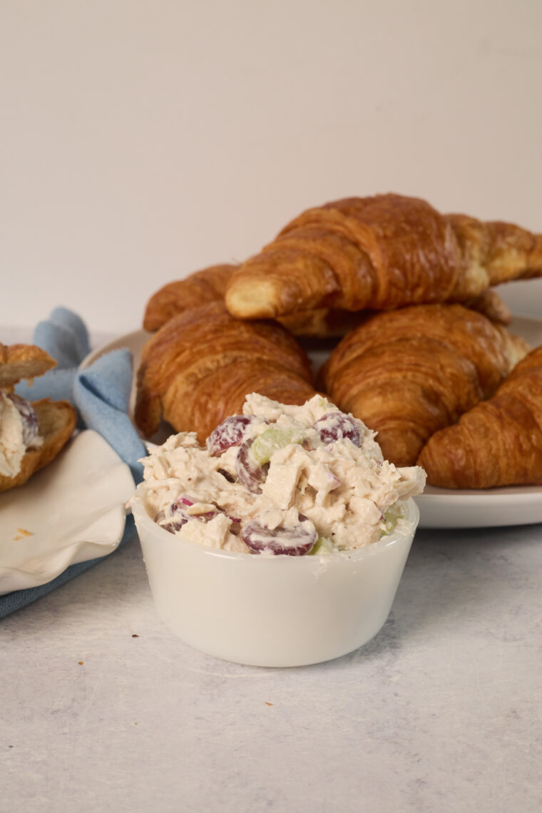 A white bowl of chicken salad with grapes and celery sits by croissants on a light surface, ready for any tasty meal.
