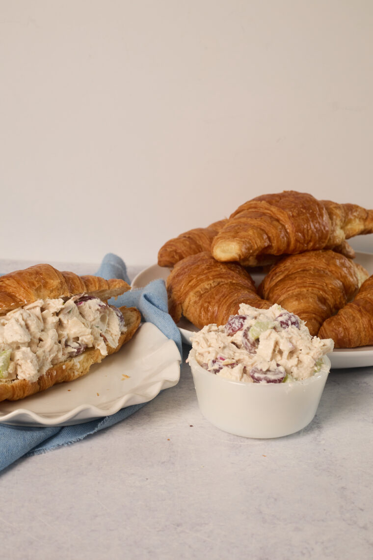 A plate of croissants, a chicken salad croissant with grapes and almonds, plus extra salad on a white table with a blue napkin.