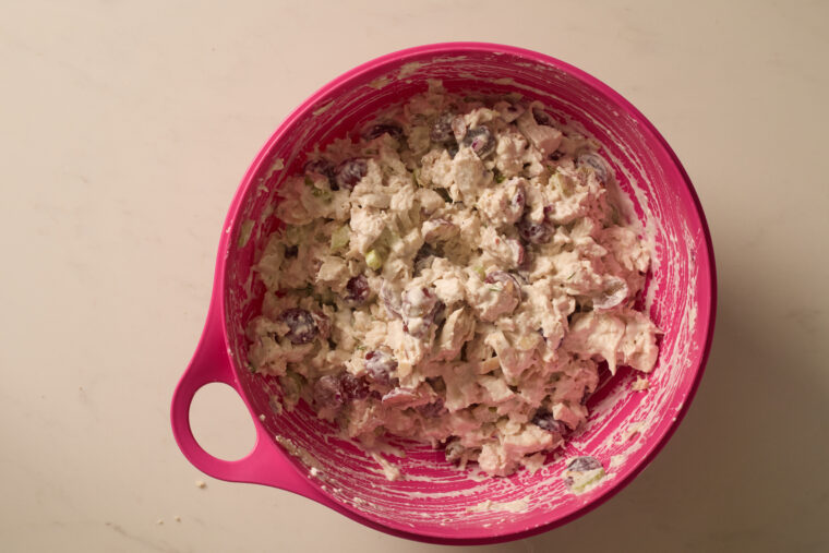 A pink bowl packed with creamy chicken salad, loaded with grapes, almonds, and veggies, sits on a light counter.