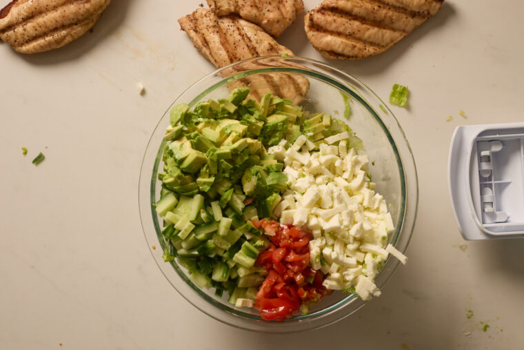 A glass bowl of salad packed with avocado, cucumber, tomato, and feta sits on the counter with some grilled chicken on the side.