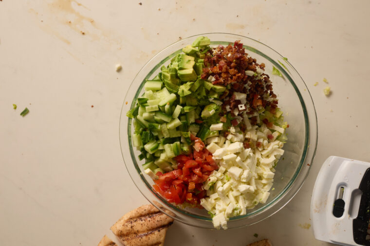 A glass bowl filled with chopped salad&mdash;cucumber, tomato, feta, bacon&mdash;sits by pita bread on a white counter.