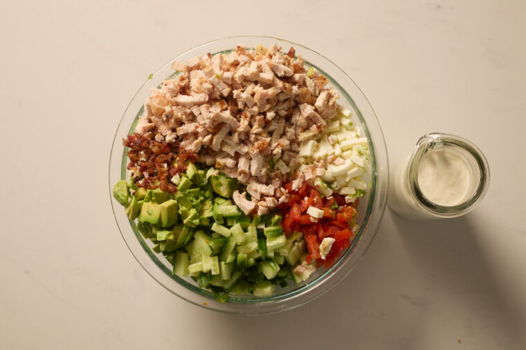 A big glass bowl packed with chopped salad&mdash;chicken, bacon, avo, cukes, tomato, lettuce&mdash;plus a jar of creamy dressing nearby.