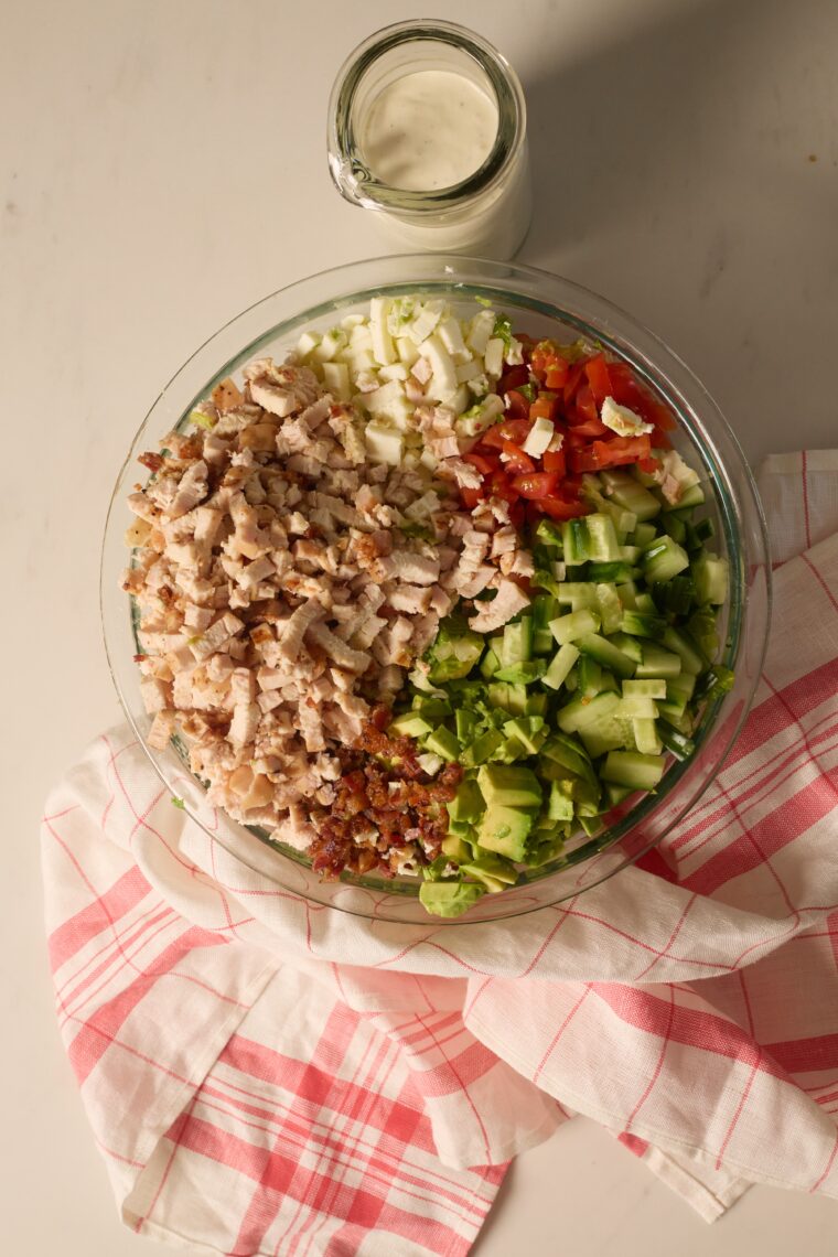 A glass bowl packed with chopped chicken, veggies, and bacon sits on a checkered towel by creamy dressing.