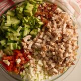 A glass bowl with a colorful chopped salad&mdash;chicken, avocado, cucumber, tomato, egg, and bacon&mdash;all sectioned off. Checkered cloth behind.