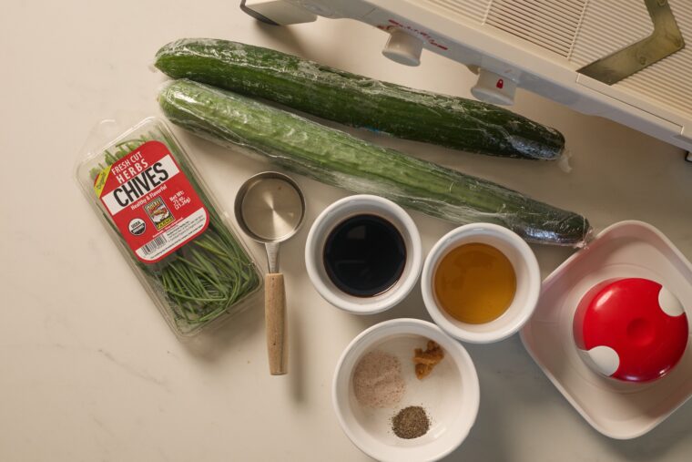 All the fixings for an Asian cucumber salad&mdash;wrapped cukes, chives, sauces, spices, and a mandoline&mdash;are on a white counter.