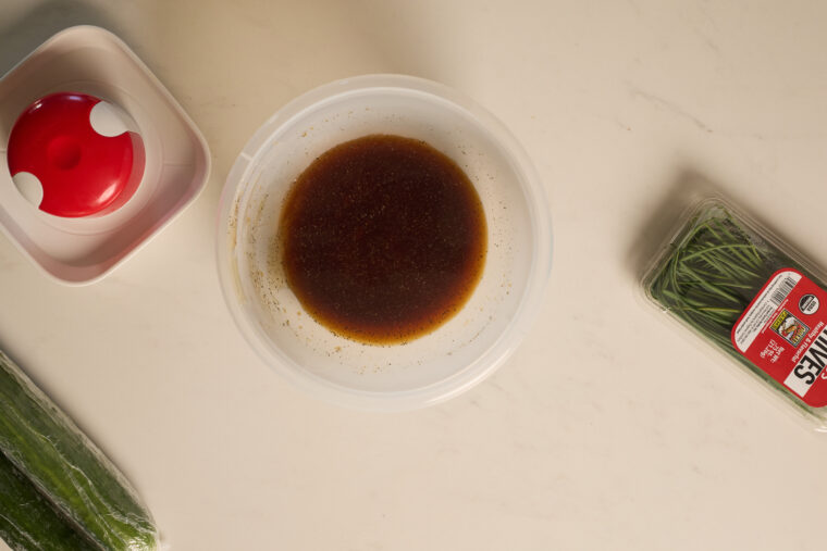 On a white counter: a plastic tub of dark sauce for Asian cucumber salad, some cucumbers, chives, and a red-and-white tool.