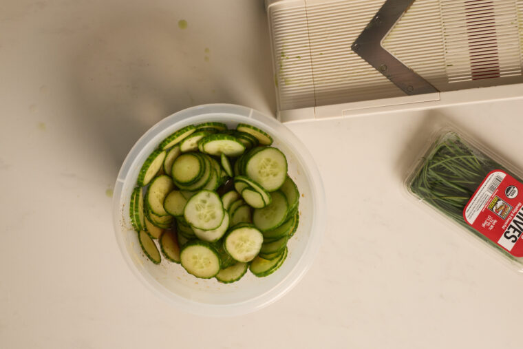 Sliced cucumbers in a bowl chill on a white counter with a mandoline and chives, prepped for a cool cucumber salad.