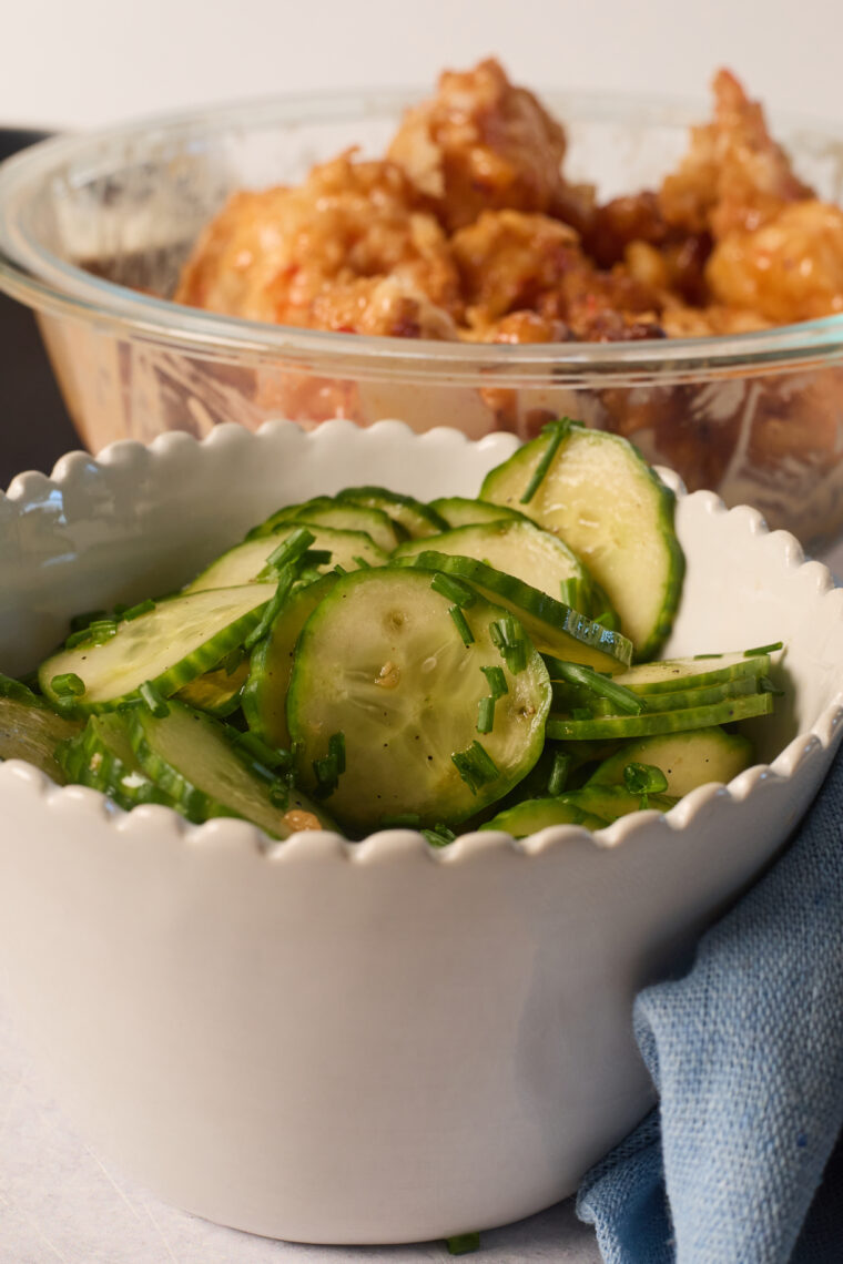 A white bowl of Asian cucumber salad topped with fresh herbs sits up front, with fried chicken pieces behind in a glass bowl.