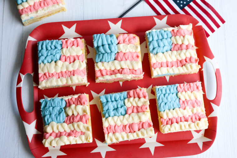 Six flag sugar cookie bars with red, white, and blue frosting sit on a starry red tray beside a little American flag.