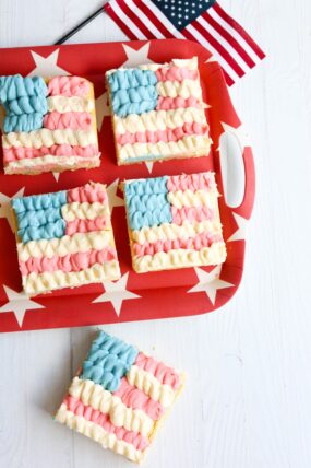 Four sugar cookie bars decorated like American flags sit on a red tray with stars. One bar is off to the side, next to a tiny flag.