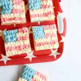 Four sugar cookie bars decorated like American flags sit on a red tray with stars. One bar is off to the side, next to a tiny flag.