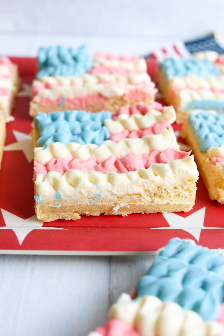 A sugar cookie bar with red, white, and blue frosting like the American flag sits on a red plate covered in white stars.