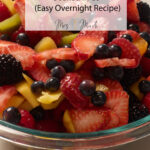 Glass bowl with strawberries, blueberries, blackberries, raspberries & mango in OJ. Text: Fresh Fruit Salad in OJ.