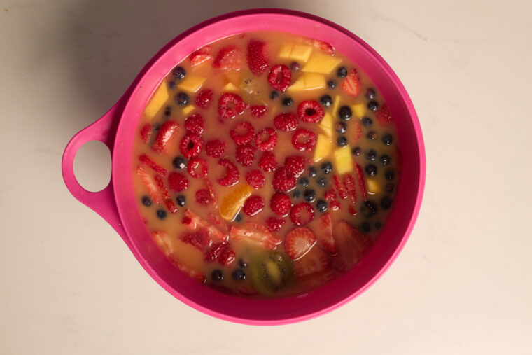 A pink bowl packed with fruit salad&mdash;chunks of fruit and berries in a yellow-orange sauce&mdash;rests on a light surface.