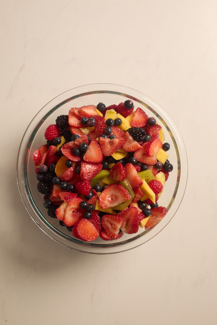 A glass bowl of simple fruit salad with strawberries, blueberries, blackberries, kiwi, raspberries, and mango on a light counter.