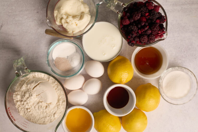Top-down look at flour, eggs, sugar, milk, butter, lemons, berries & vanilla by measuring cups on a light countertop.