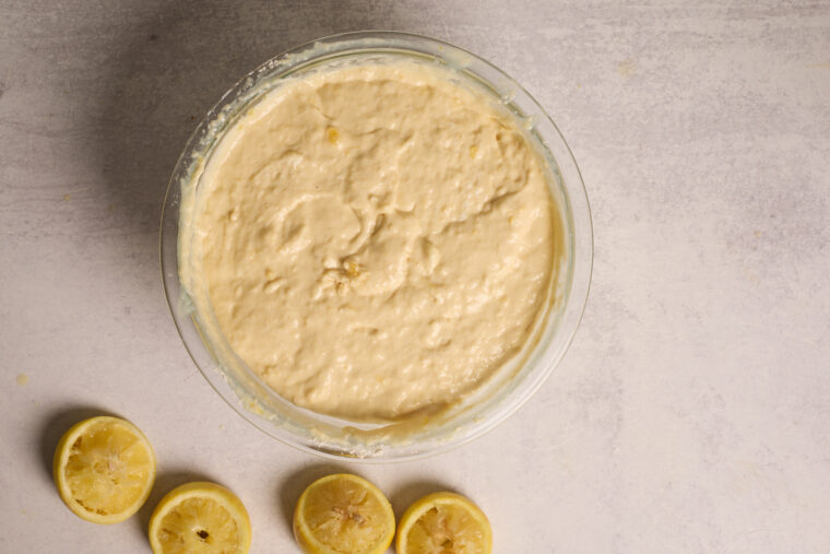 A glass bowl of cake batter sits on a light counter with three squeezed lemon halves—great for whipping up zesty pancakes.