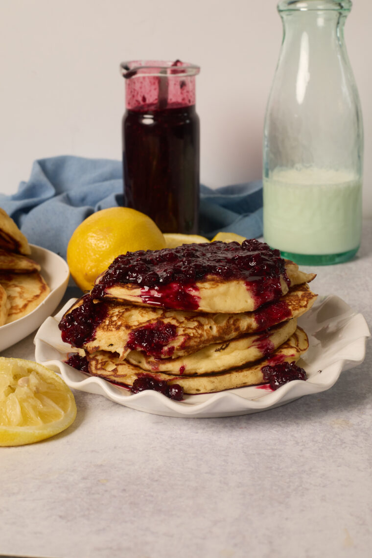 A stack of lemony pancakes with berry sauce sits on a plate, with a lemon, jam jar, and milk bottle behind them.