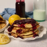 A stack of lemony pancakes with berry sauce sits on a plate, with a lemon, jam jar, and milk bottle behind them.