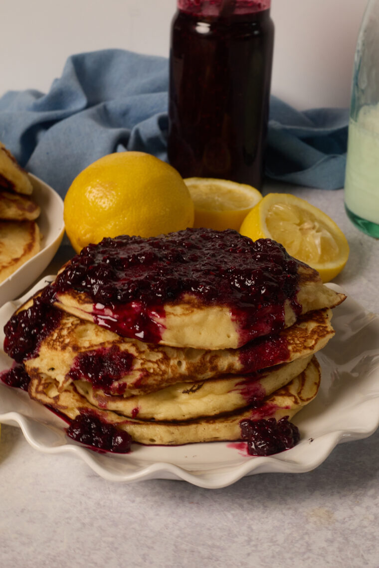 A plate of zesty pancakes piled high with berry sauce, with lemons, a sauce jar, and a bottle hanging out in the back.