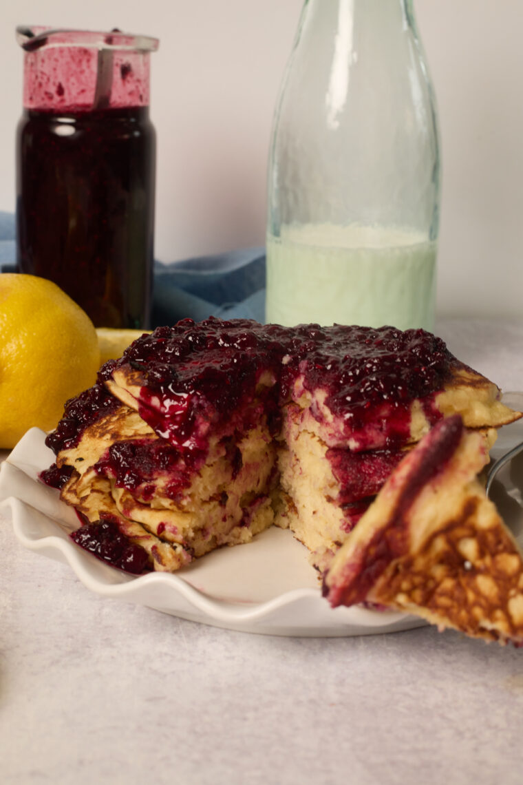 A stack of lemon ricotta pancakes with berry sauce on a plate, one slice missing. Jar of sauce, lemon, and milk bottle behind.