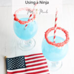 Two blue slushi mocktails with red sugar rims and striped straws sit by a tiny American flag. Text says: Patriotic Slushi Mocktail Using a Ninja.