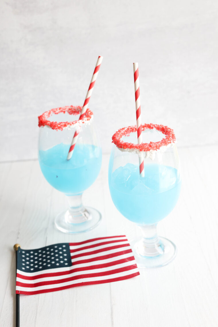 Two blue slushies with red sugar rims and striped straws chill on a white table beside a tiny American flag.