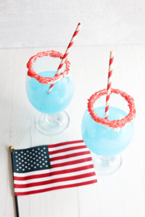 Two blue slushies with red sugar rims and striped straws sit next to a tiny American flag on a white table.