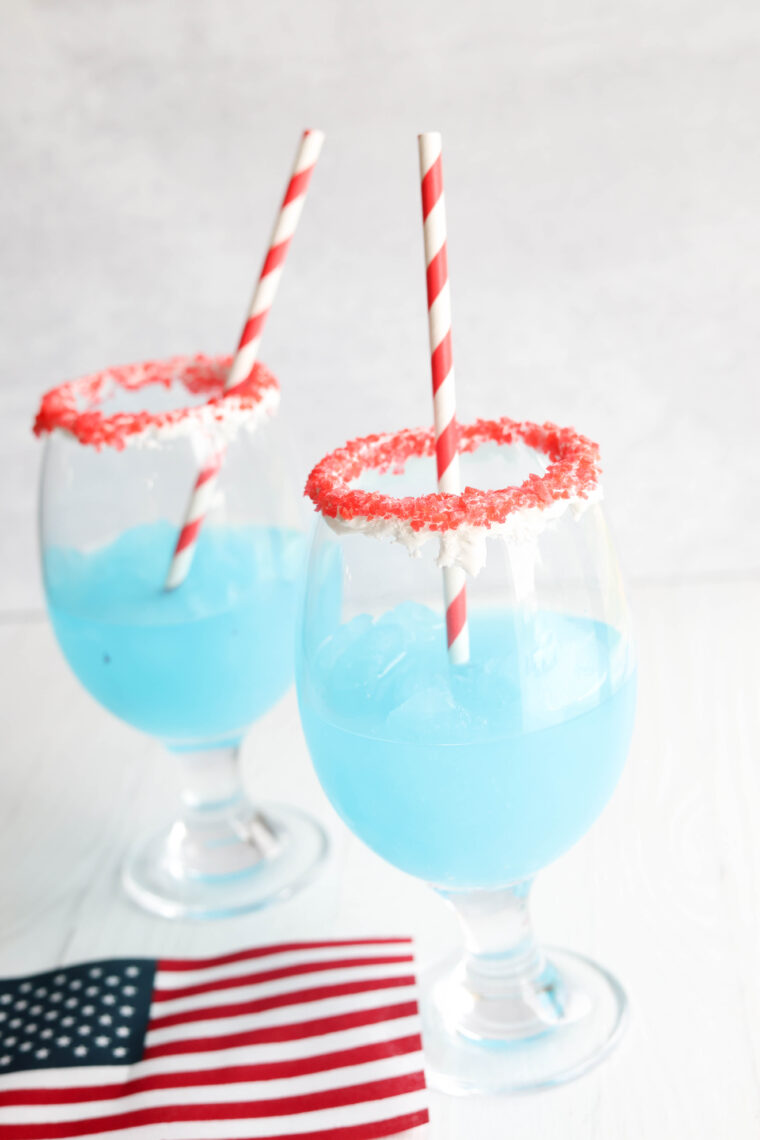 Two fancy glasses filled with blue slushies, topped with red sugar rims and striped straws. An American flag is up front.