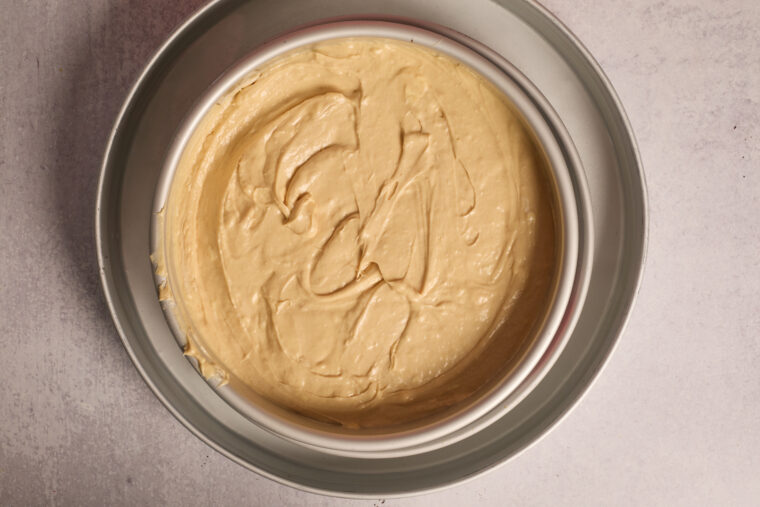 A metal cake pan with a layer of creamy peanut butter cheesecake batter rests on a light-colored counter.
