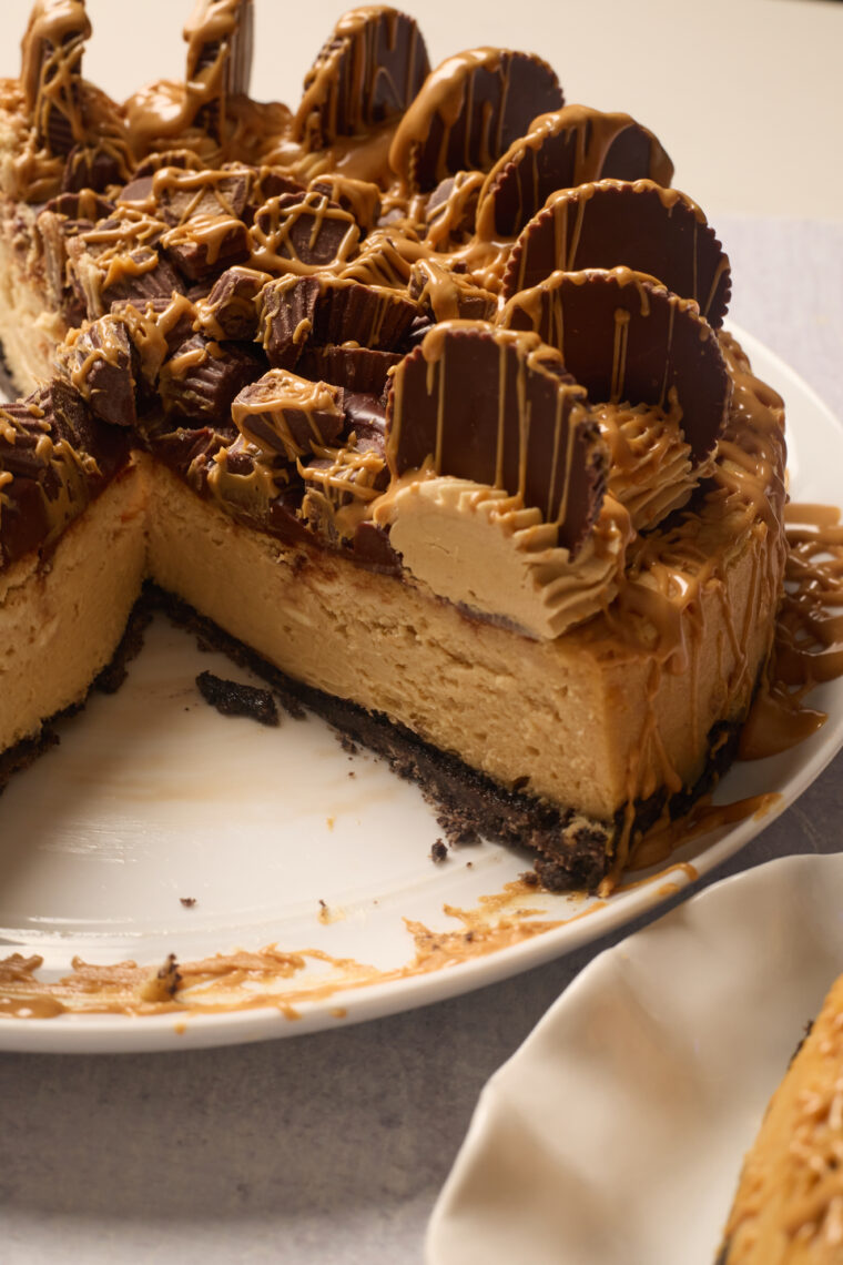 A creamy peanut butter cheesecake with a slice gone, loaded with PB cups and peanut butter drizzle on a white plate.
