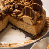 A creamy peanut butter cheesecake with a slice gone, loaded with PB cups and peanut butter drizzle on a white plate.