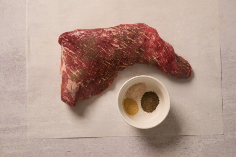 A raw tri-tip steak sits on parchment paper by a bowl with salt, pepper, and garlic powder—all ready to get cooking.