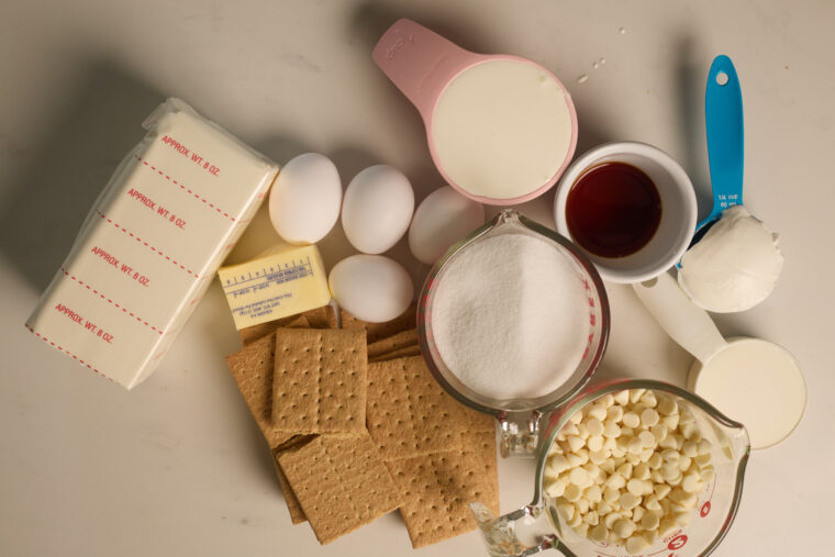 All the cheesecake basics are spread out&mdash;cream cheese, eggs, butter, graham crackers, sugar, vanilla, white chocolate chips, milk.