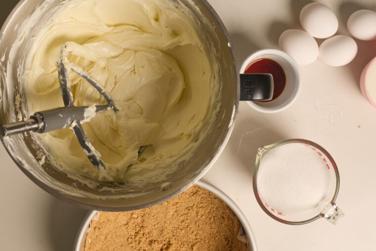 Overhead shot of creamy batter in a bowl, with eggs, sugar, vanilla, and graham crackers around&mdash;cheesecake time!.