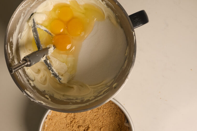 A bowl of cream cheese, sugar, and eggs being mixed, next to graham crumbs&mdash;cheesecake magic in the making!.