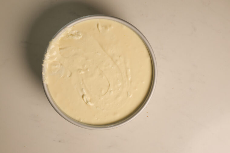 A round pan with smooth, white chocolate cheesecake batter sits on a pale countertop, ready to be baked.