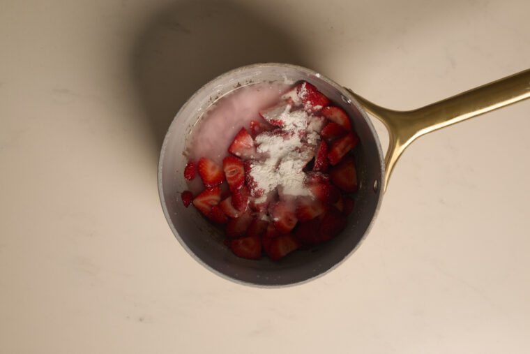 Chopped strawberries, cream, and white powder mix in a saucepan on a light counter&mdash;ready for cheesecake or sweet treats.