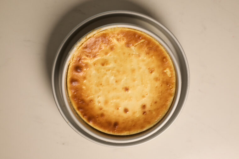 A smooth, baked cheesecake with a golden top rests in a round metal pan on a pale-colored counter.