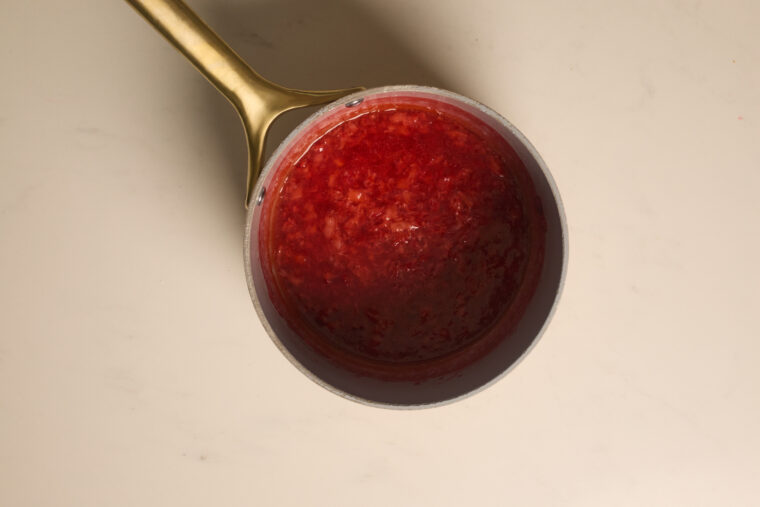 A gold-handled saucepan holds a creamy red, strawberry cheesecake-like sauce on a light countertop.